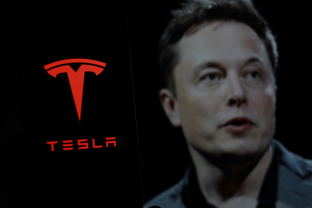 Tesla logo is displayed on smartphone screen With CEO Elon Musk in a background. — Photo by MuhammadAlimaki