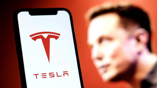 The Tesla logo is displayed on smartphone screen With CEO Elon Musk in a background — Photo by MuhammadAlimaki