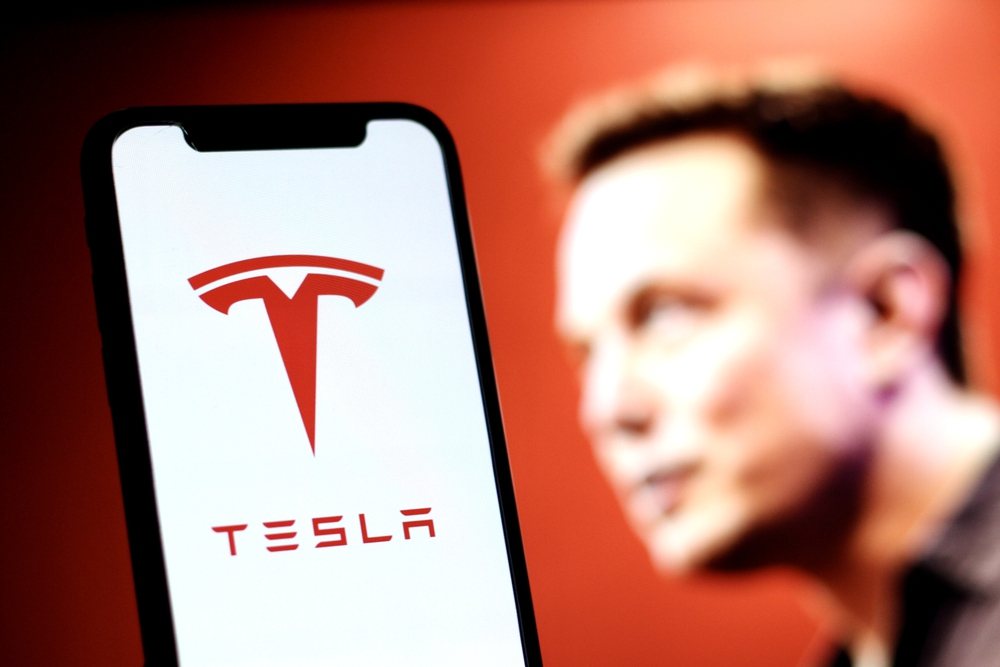 The Tesla logo is displayed on smartphone screen With CEO Elon Musk in a background — Photo by MuhammadAlimaki