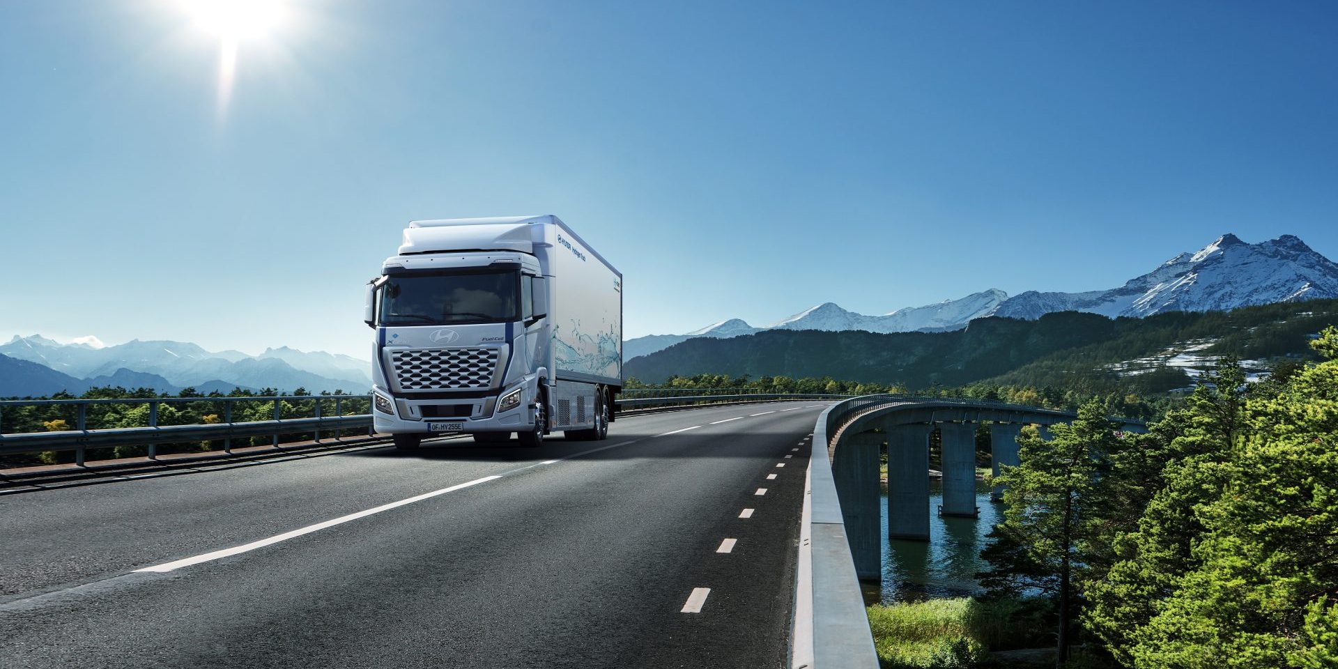 A red truck travels on a snowy road, set against a stunning backdrop of snow-covered mountains and wind turbines.