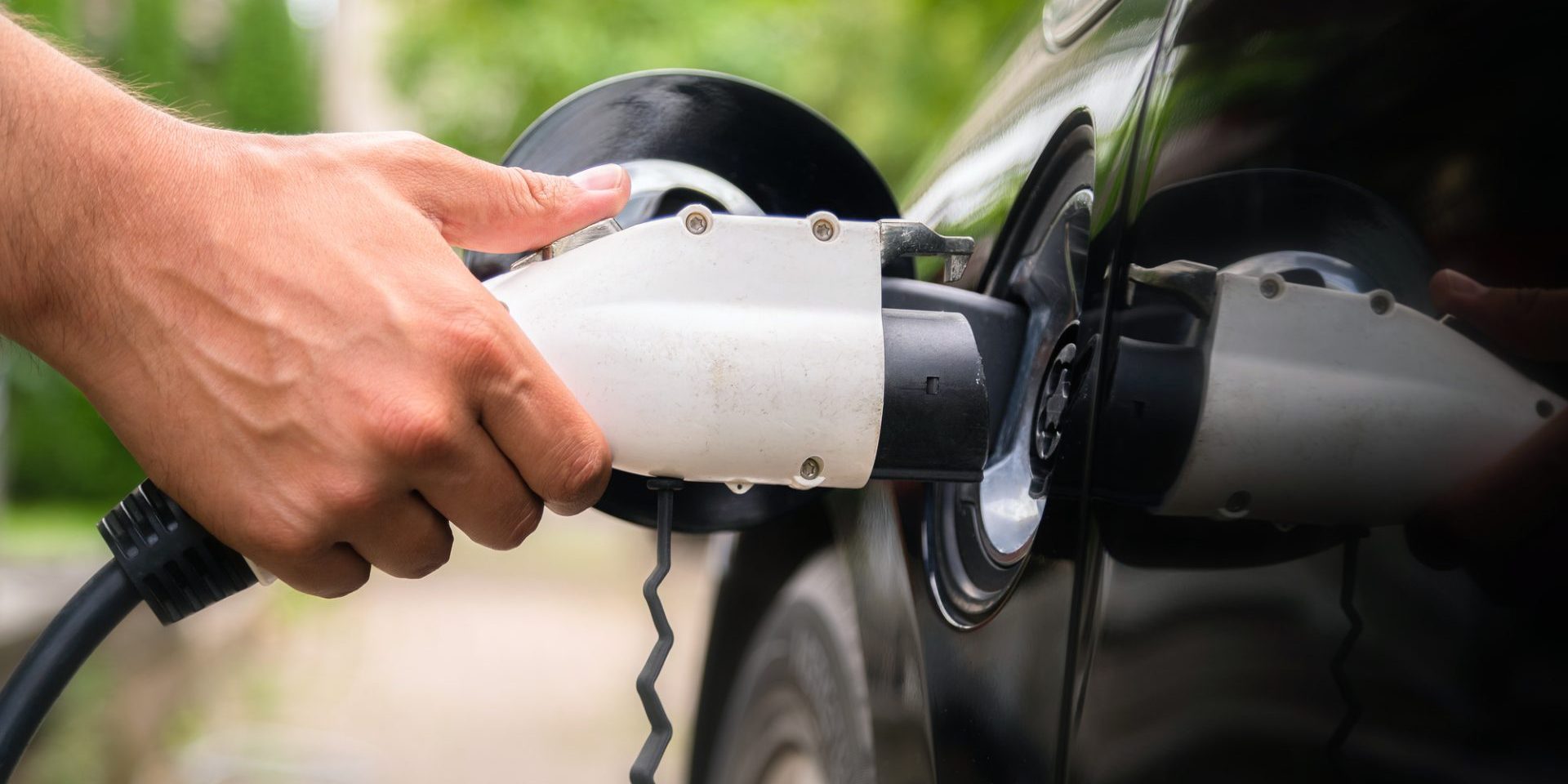 Mans hand inserting charger plug into electric car in green environment background. New energy vehicle, NEV is being loaded with electricity power. Ecology, modern day cars