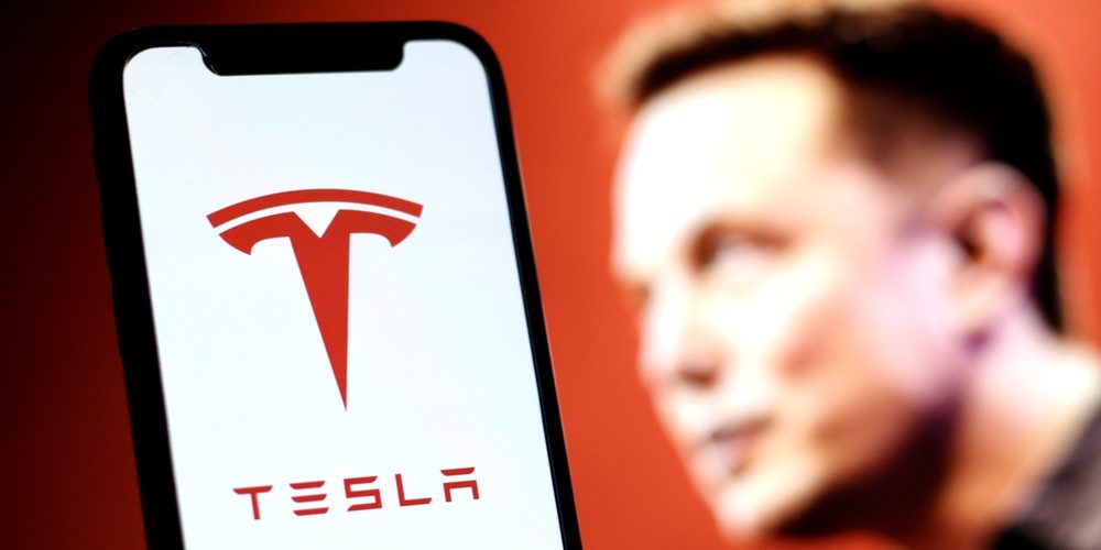The Tesla logo is displayed on smartphone screen With CEO Elon Musk in a background — Photo by MuhammadAlimaki